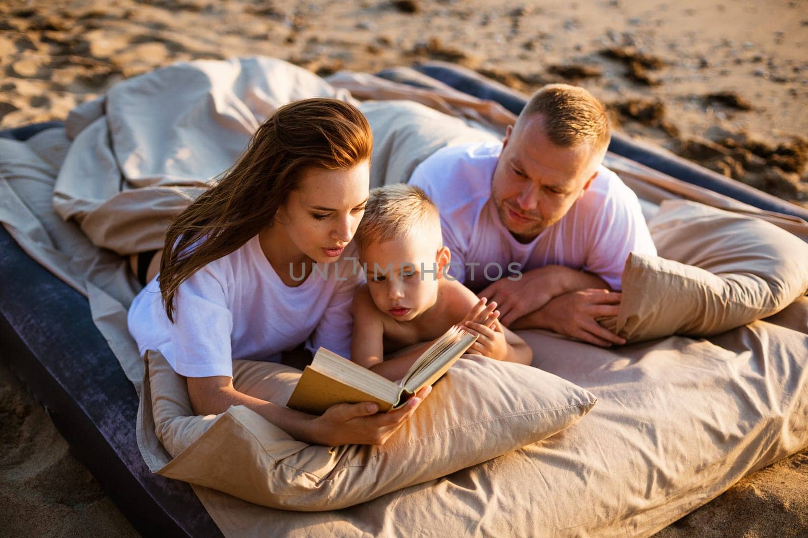 Mom is reading a book to her son while lying on the bed. Family vacation by the sea. Happy young caucasian family resting together at sunset