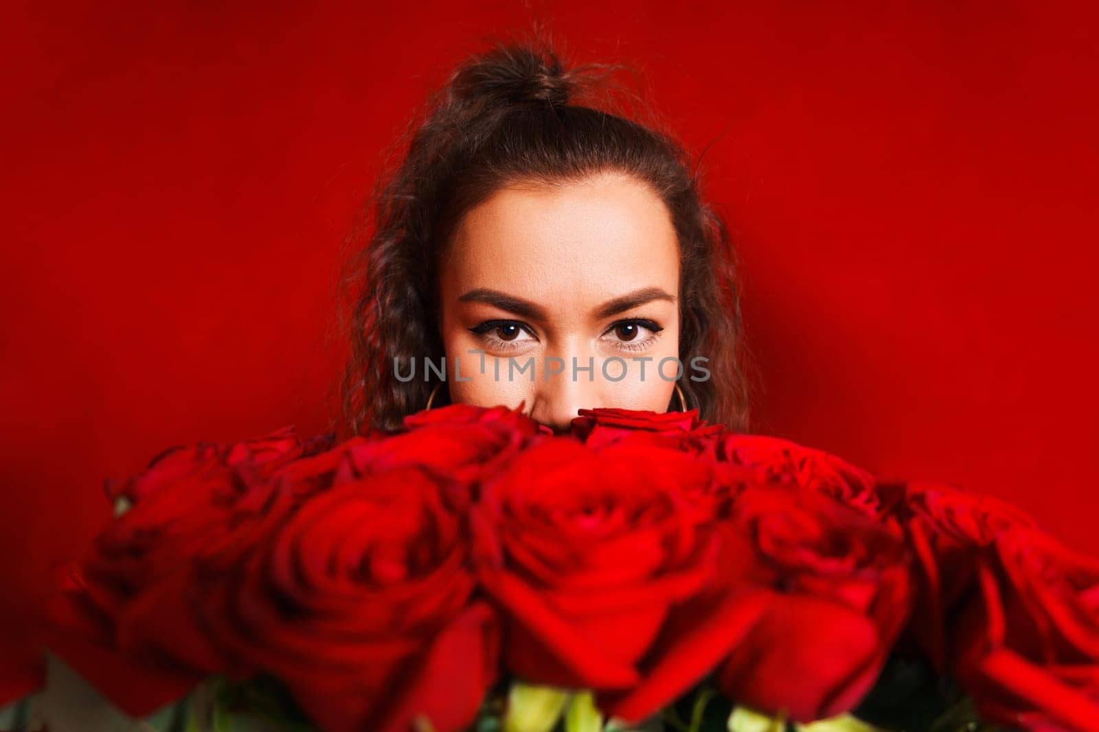 Portrait of girl peeking out from behind bouquet of flowers on red background. Portrait of girl peeks out from behind bouquet of flowers on red background. Young woman holding large bouquet red roses