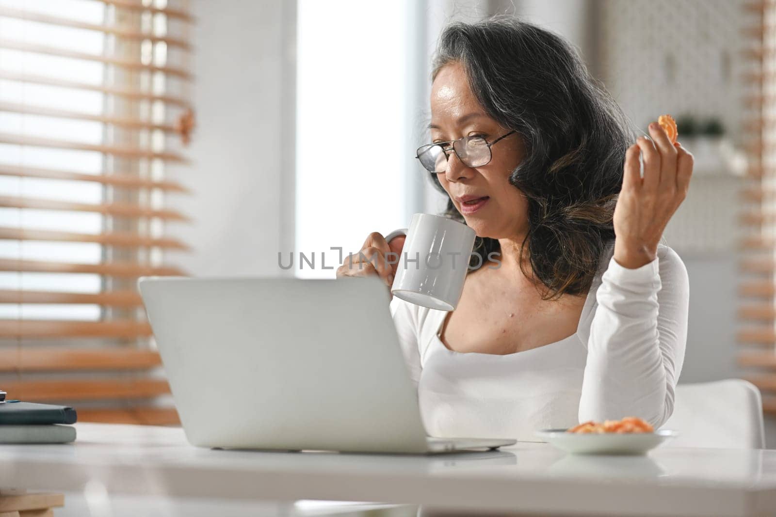 Portrait of beautiful middle aged woman drinking hot coffee and looking at laptop screen.