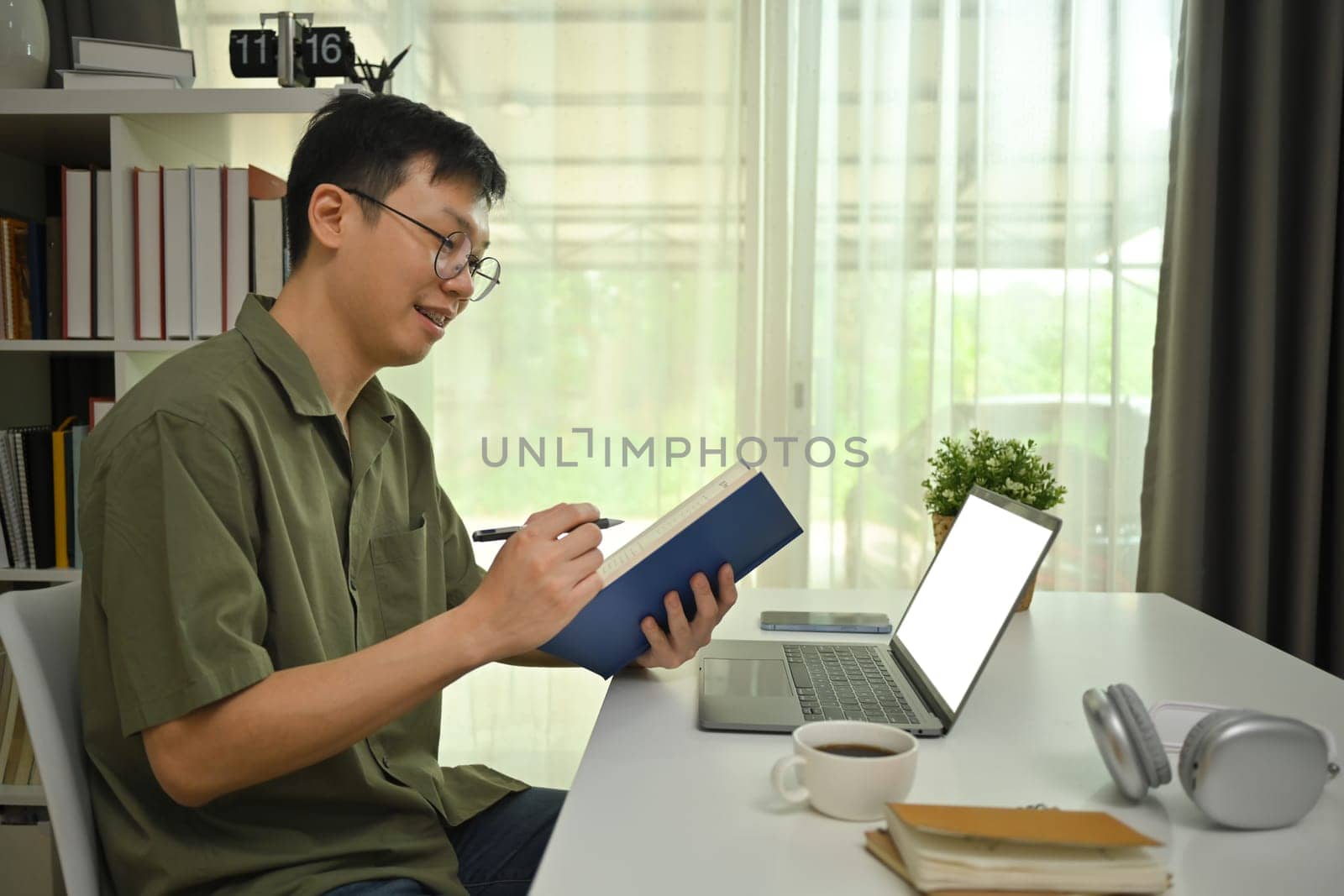 Young man in glasses working remotely from home office with laptop and making notes on notebook by prathanchorruangsak