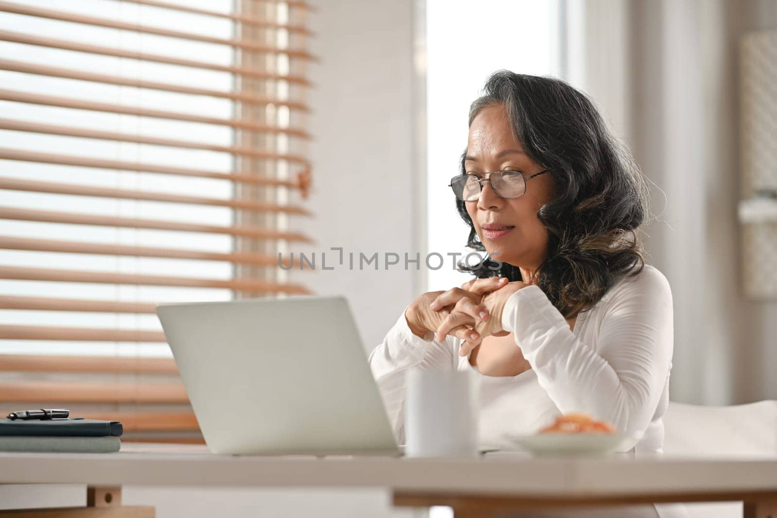 Middle aged woman in glasses watching online webinar on laptop during remote working from home.
