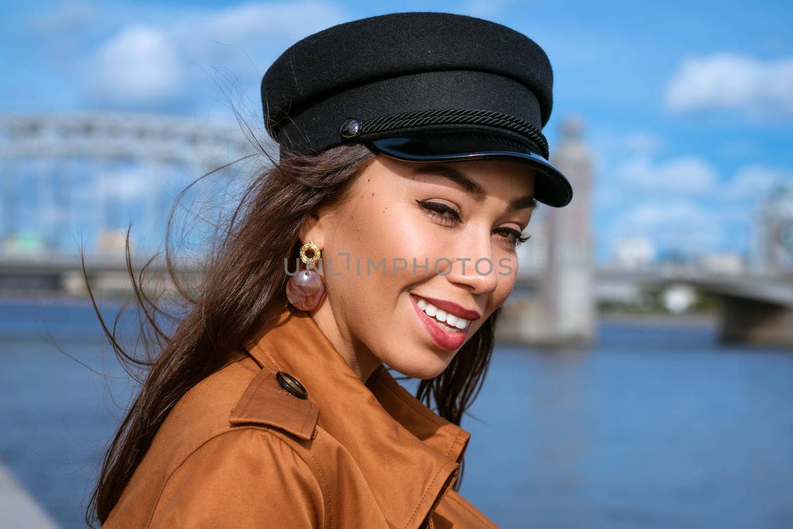 portrait of a happy Caucasian woman on the embankment of the river in a black cap and brown jacket on a sunny day