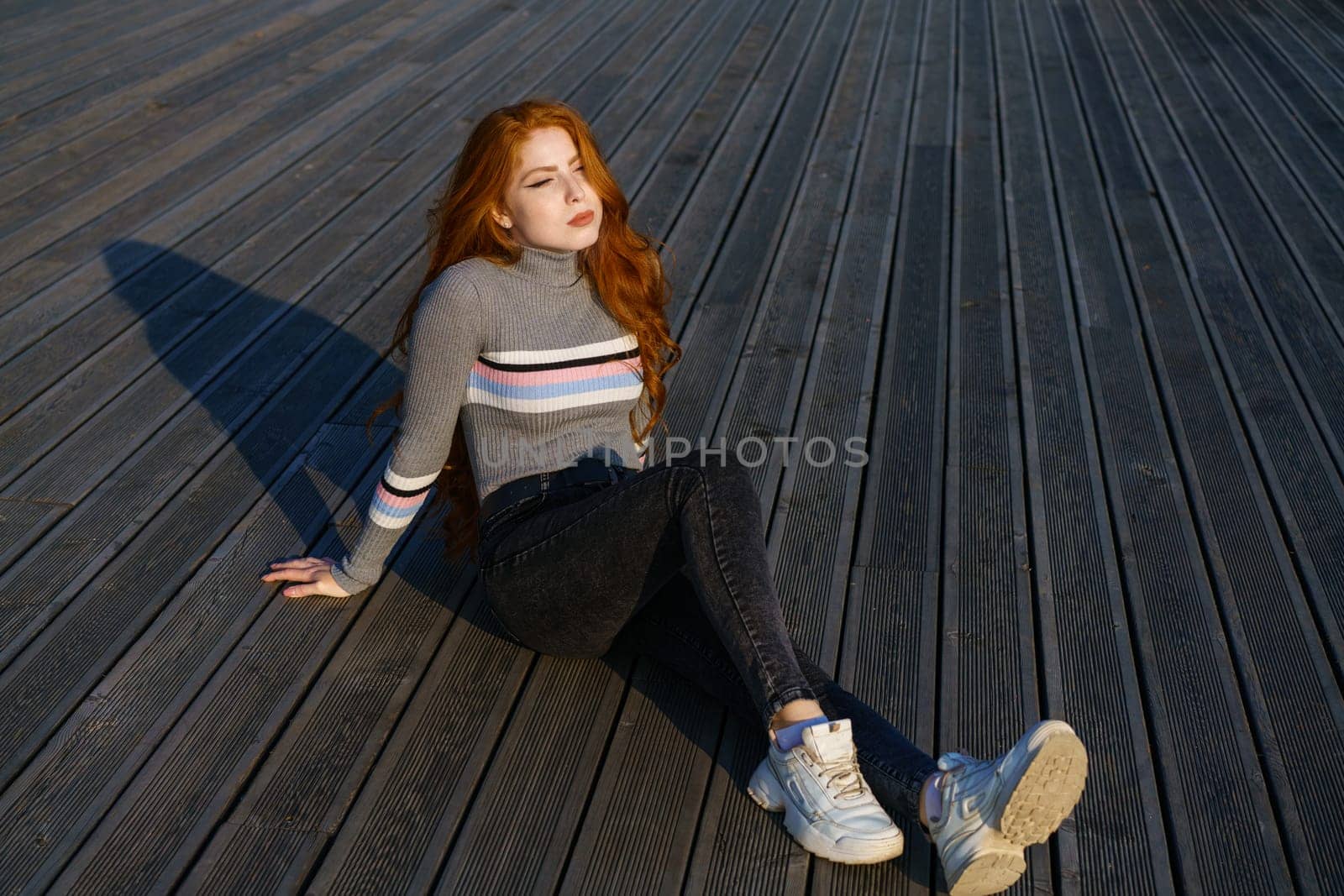 sad redhead girl sits on a wooden background in the park on a sunny day alone. Young woman alone. Sadness and depression concept