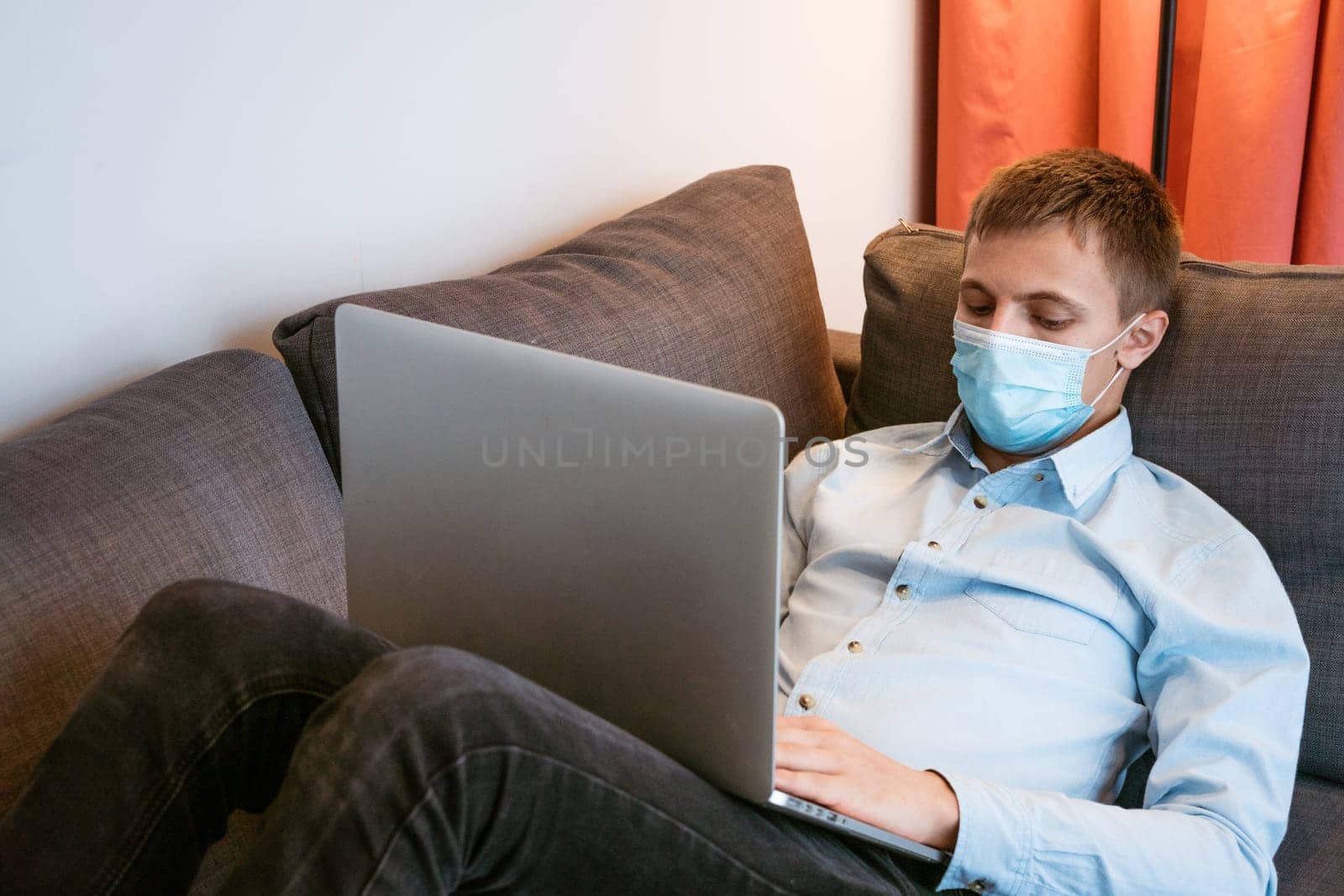 young man in protective mask with laptop on sofa by EkaterinaPereslavtseva