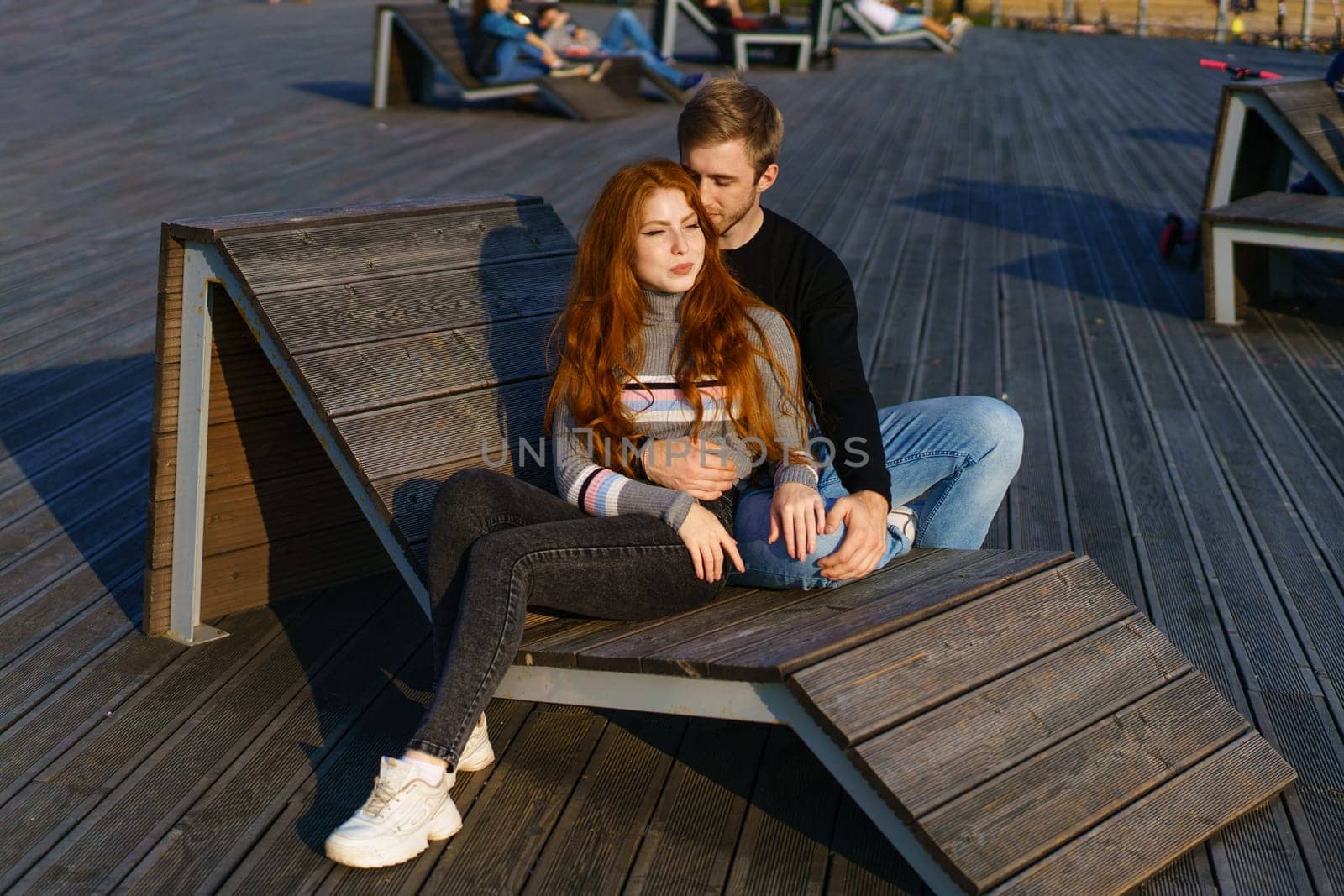 young couple of guy and girl with red hair of Caucasian appearance, in casual clothes, on a sunny day sitting in the park on a wooden bench in an embrace, happy relationship between a man and a woman