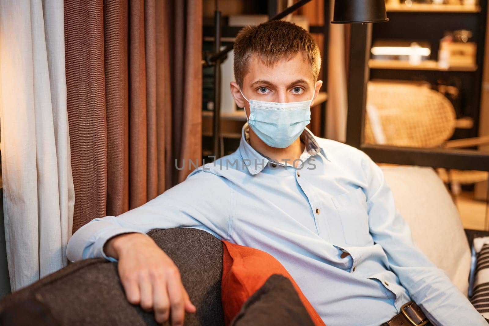 a young guy of Caucasian ethnicity in a shirt and a protective mask sits on the couch alone at home. Concept of being at home during global quarantine