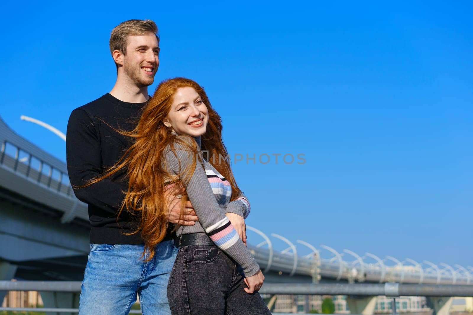 happy young couple man and woman with long red hair, stand against the background of a blue sky and a bridge in casual clothes and smile. Cheerful guy and Caucasian girl on a sunny day