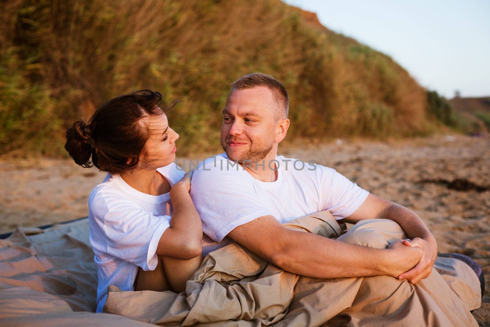 Young couple wrapped in a blanket outdoors. Happy couple in love lying and rotating in bed by the sea at sunset. Happy caucasian couple of husband and wife wrapped in a blanket outdoors.