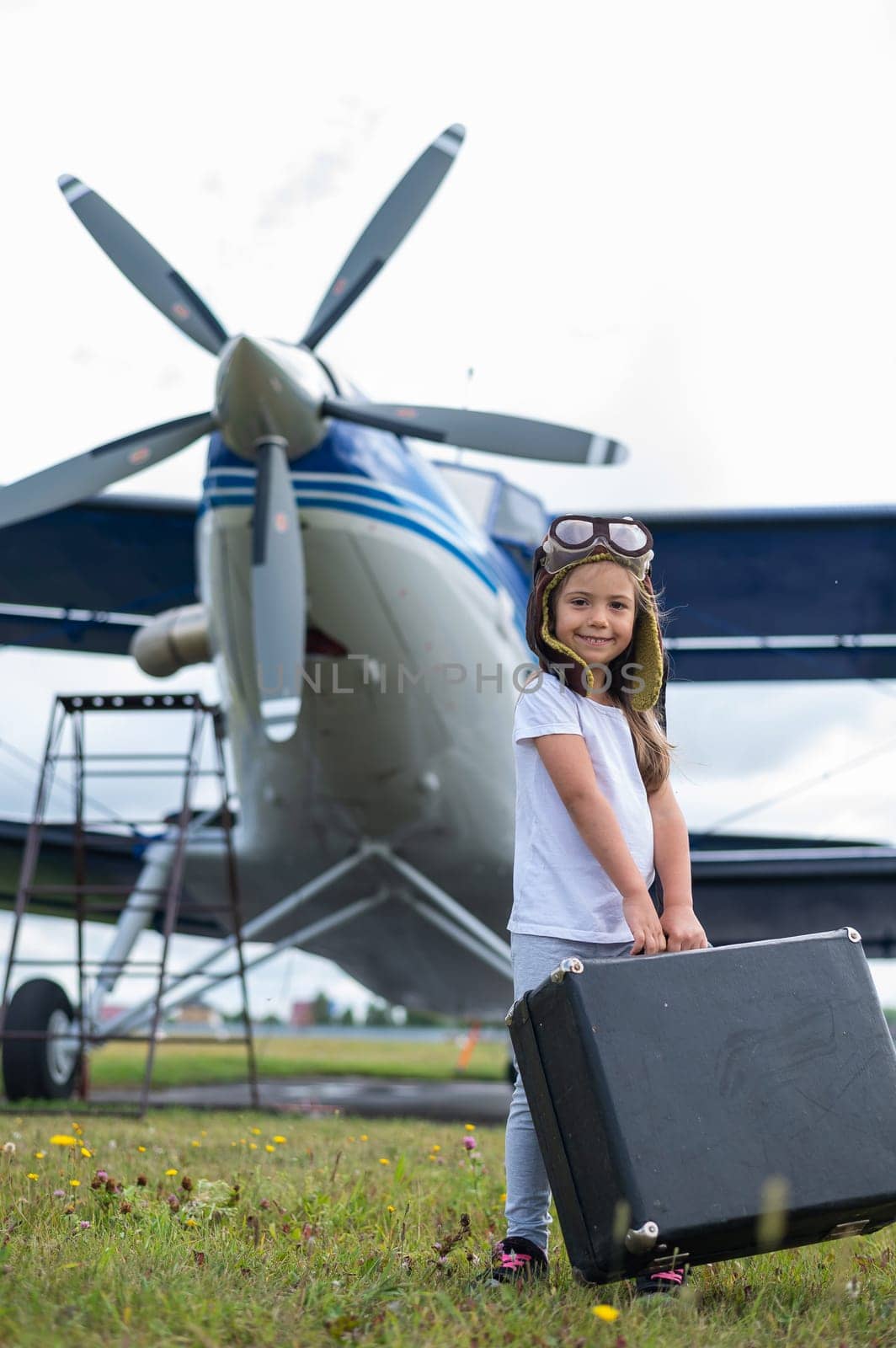 A little girl in a pilot's costume carries a retro suitcase and walks along the airfield. A child in a hat and glasses is going on a trip by plane. by mrwed54