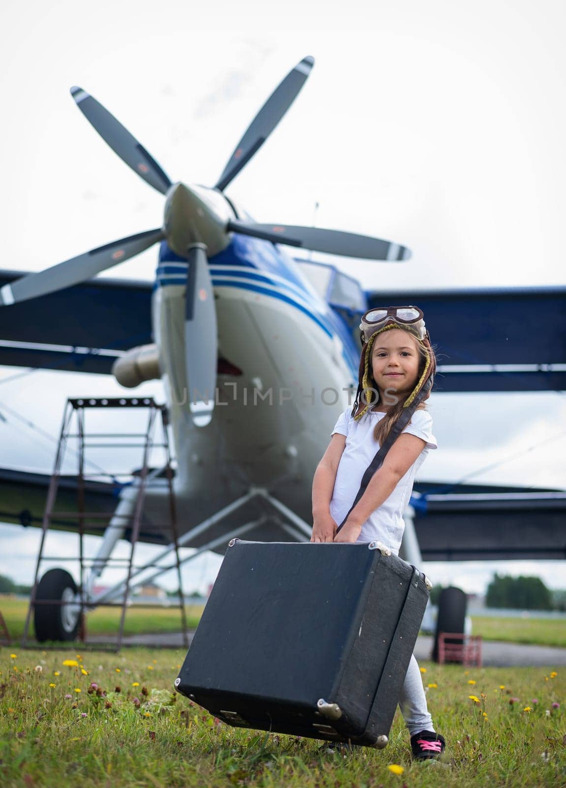 A little girl in a pilot's costume carries a retro suitcase and walks along the airfield. A child in a hat and glasses is going on a trip by plane. by mrwed54