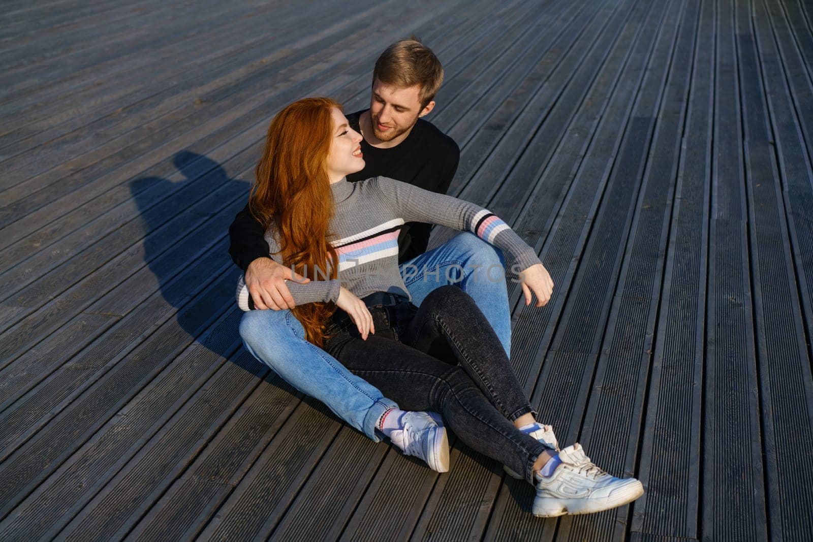 young couple of guy and girl with red hair of Caucasian appearance, in casual clothes, on a sunny day sitting in the park on a wooden bench in an embrace, happy relationship between a man and a woman