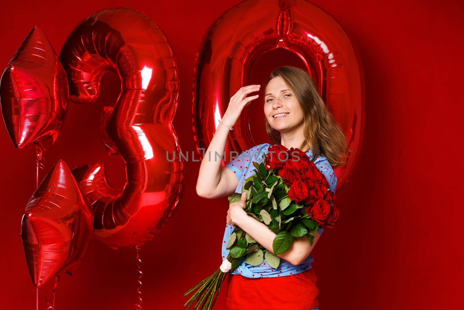 Beautiful young woman on a red background with red balloons and a bouquet of red roses. Cheerful and happy girl with bright red lipstick celebrates 30 years anniversary