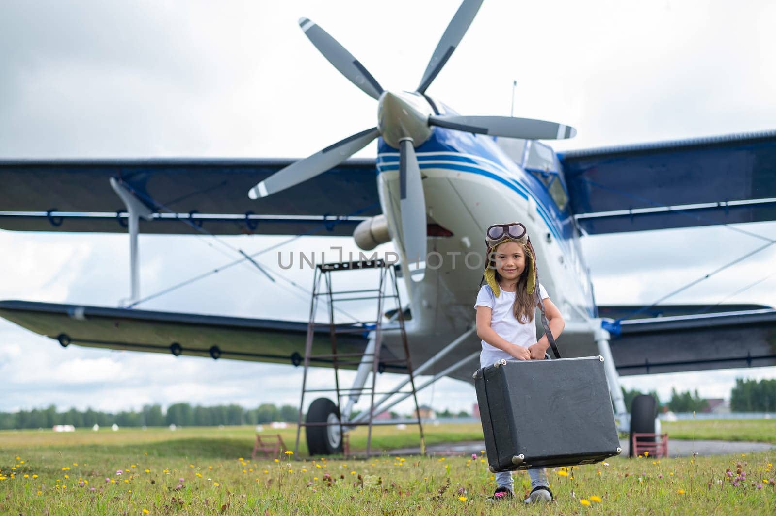 A little girl in a pilot's costume carries a retro suitcase and walks along the airfield. A child in a hat and glasses is going on a trip by plane