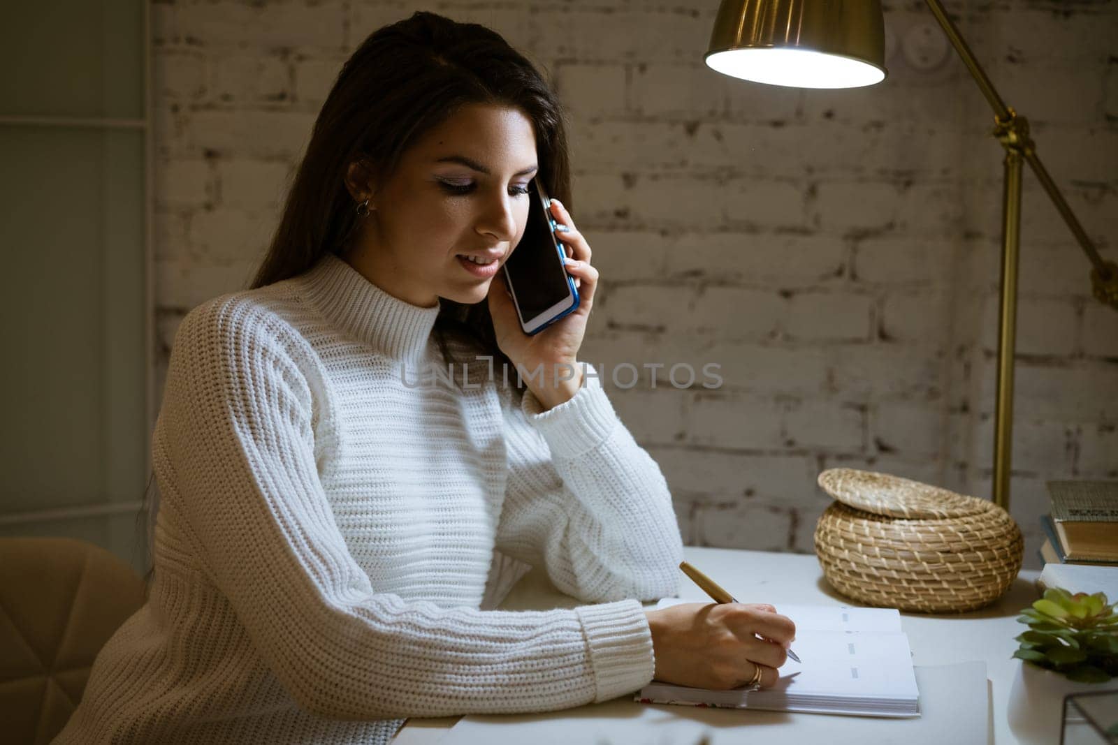 Happy woman in white sweater talking on the phone at the table