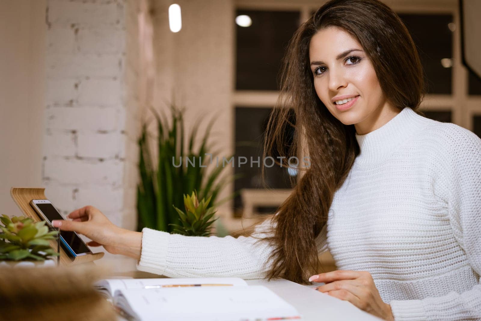 Beautiful business woman in a white sweater is working on the online communication by phone while sitting at the table