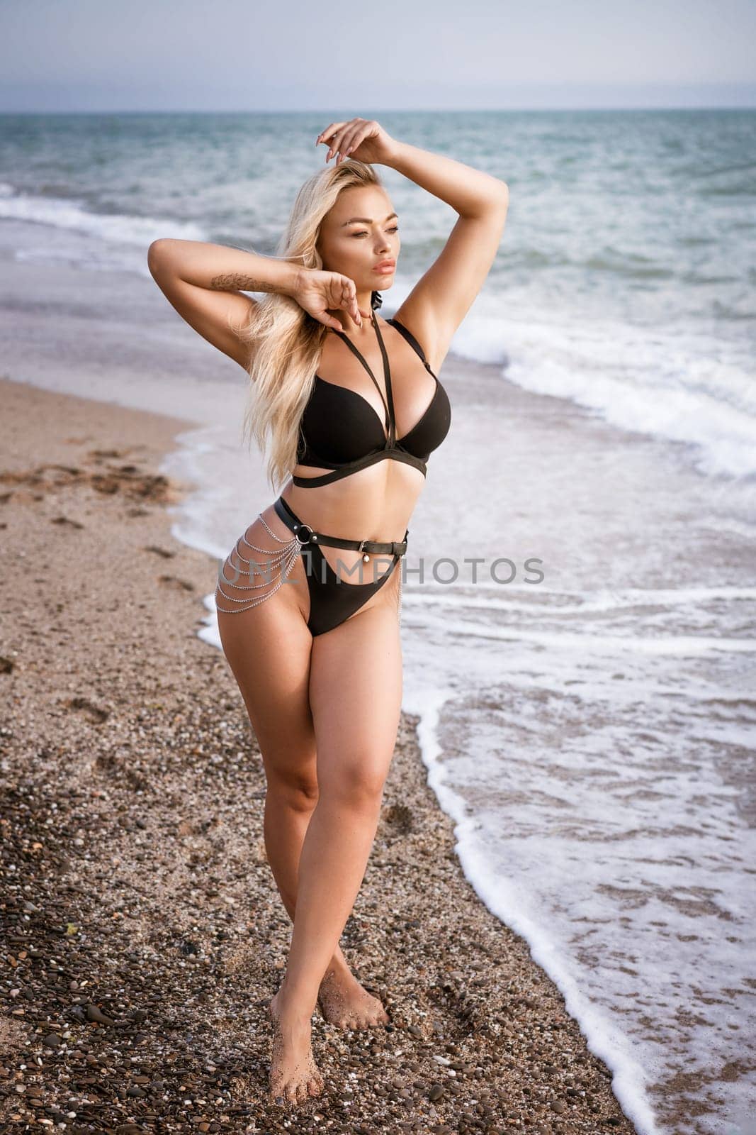 a beautiful blonde girl in an open swimsuit poses on a sandy beach, near the sea. Waves roll up on the shore, the horizon is visible in the background.