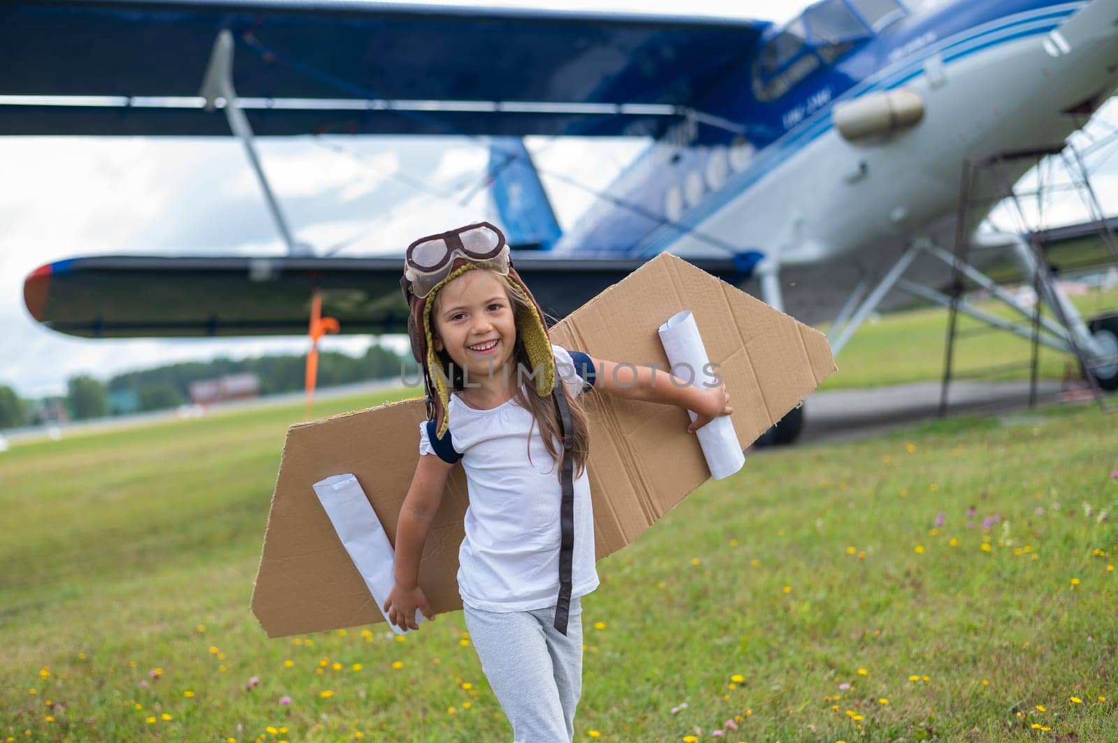 A little girl in a pilot's costume with cardboard wings runs on the lawn against the backdrop of the plane. A child in a hat and glasses dreams of flying on an airplane. by mrwed54