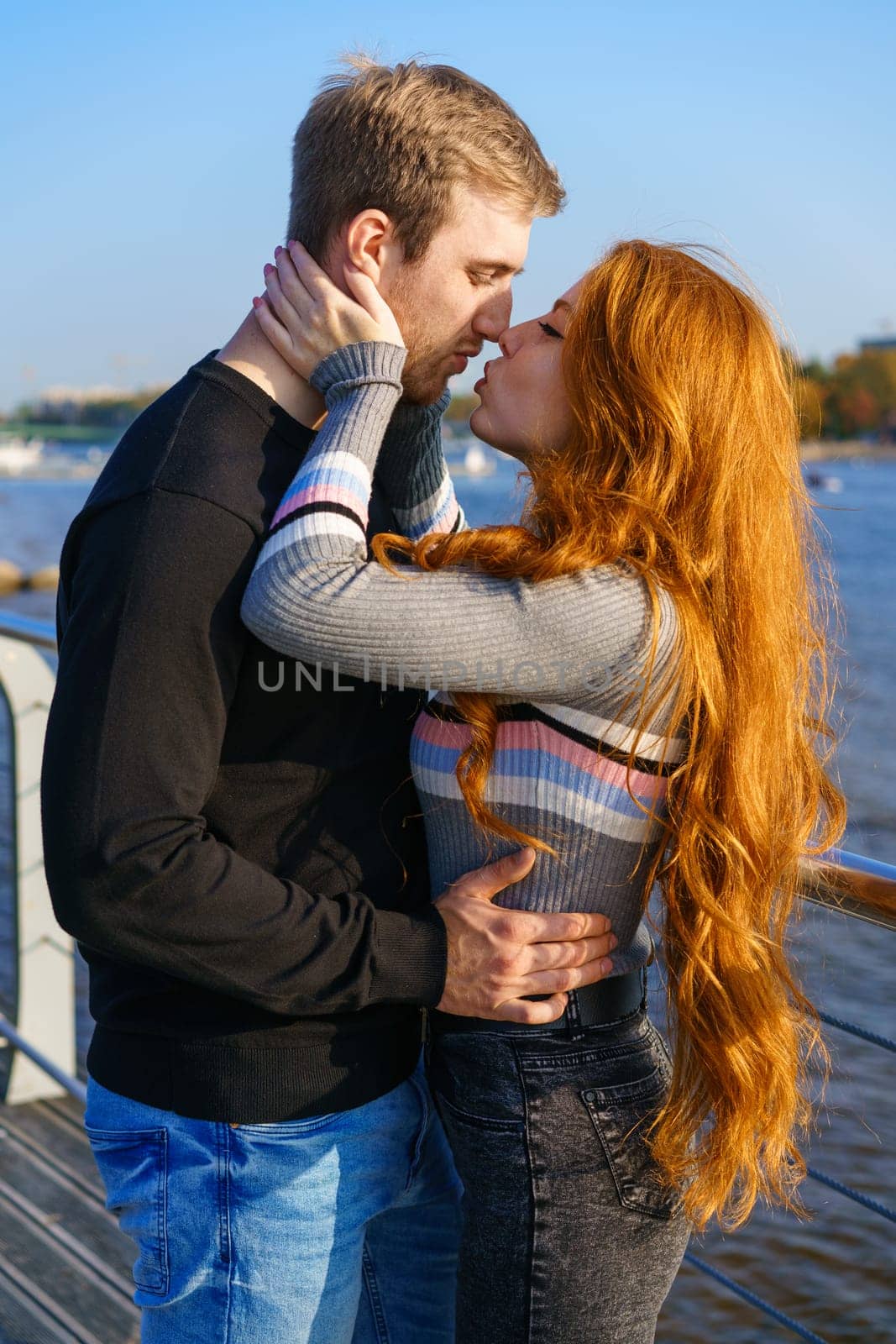 Young couple of man and woman with long red hair of Caucasian ethnicity, in casual clothes, stand on the bank of the river embracing happy on a summer day against the background of the cityscape