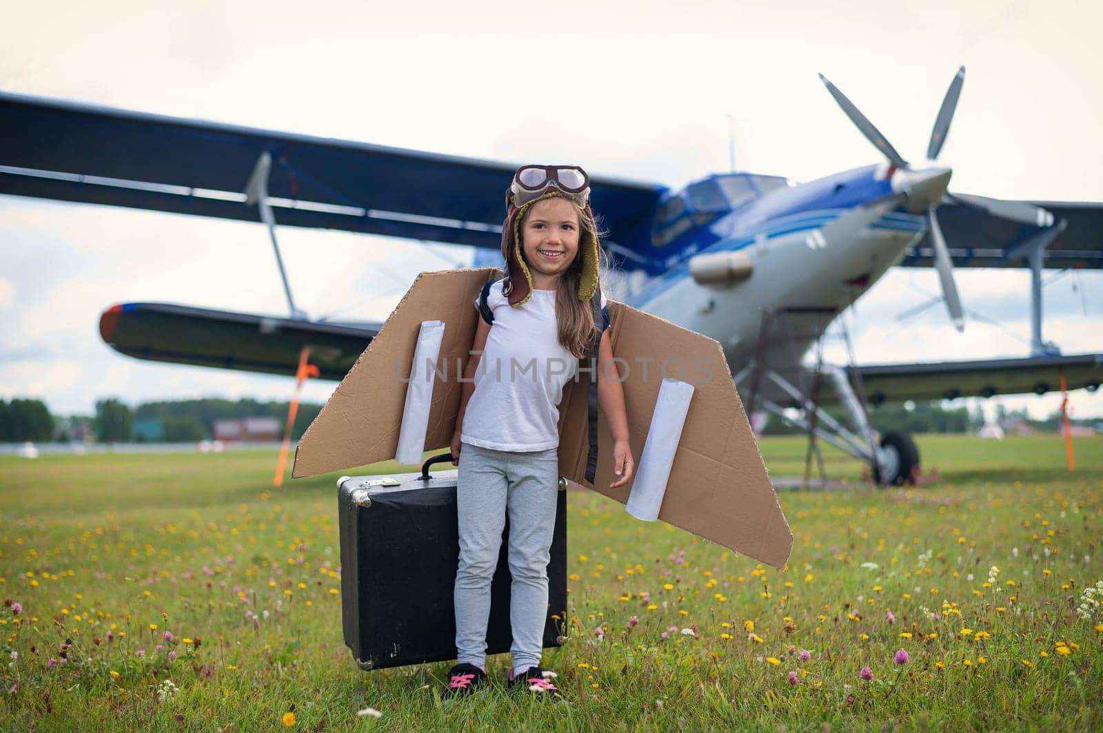 A little girl in a pilot's costume sits on a retro suitcase at the airport waiting for the departure of the flight. A child in a hat and glasses is going on a trip by plane