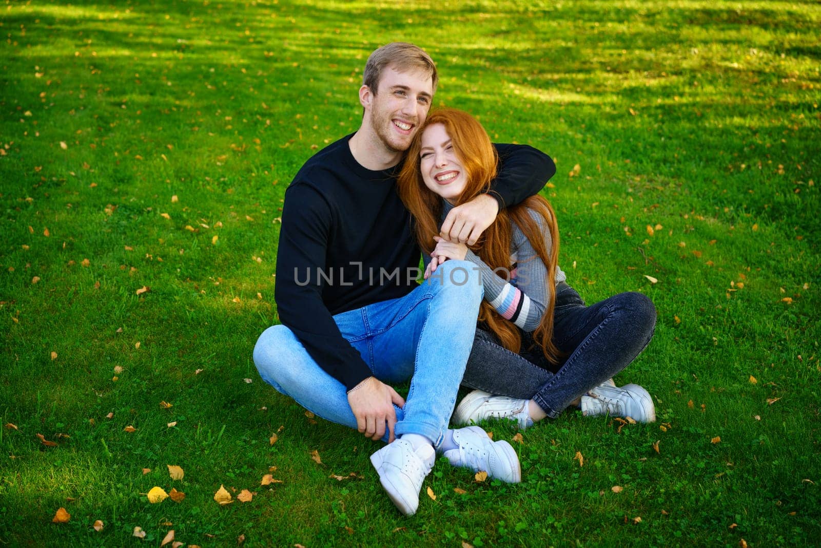 Happy young couple of Caucasian ethnicity man and woman in casual clothes sitting embracing in the park on green grass on a sunny day. Happy relationship between prana and a girl in nature