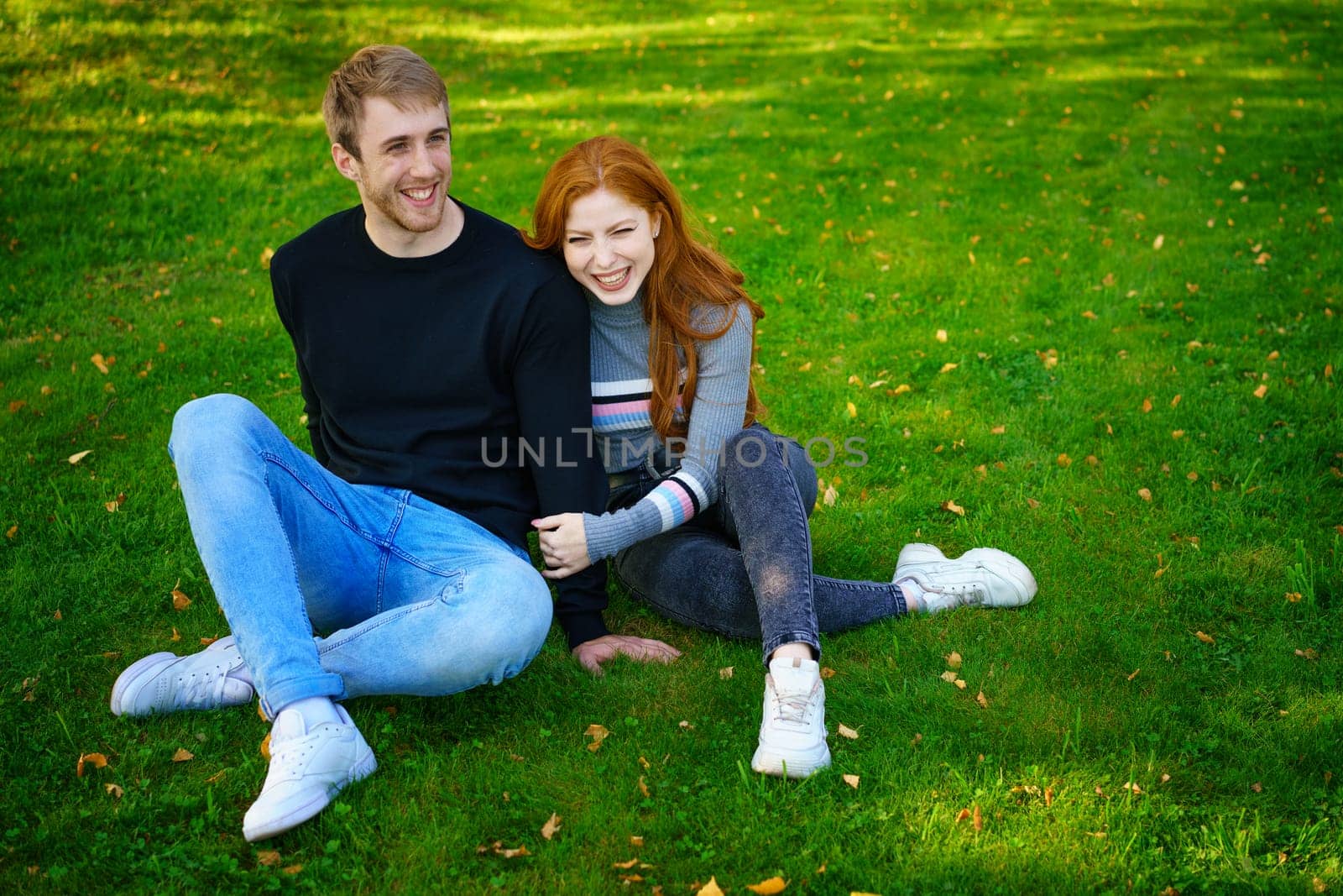 Cheerful young couple guy and red-haired girl of Caucasian ethnicity , are having fun together in the park on green grass on a sunny summer day in casual clothes
