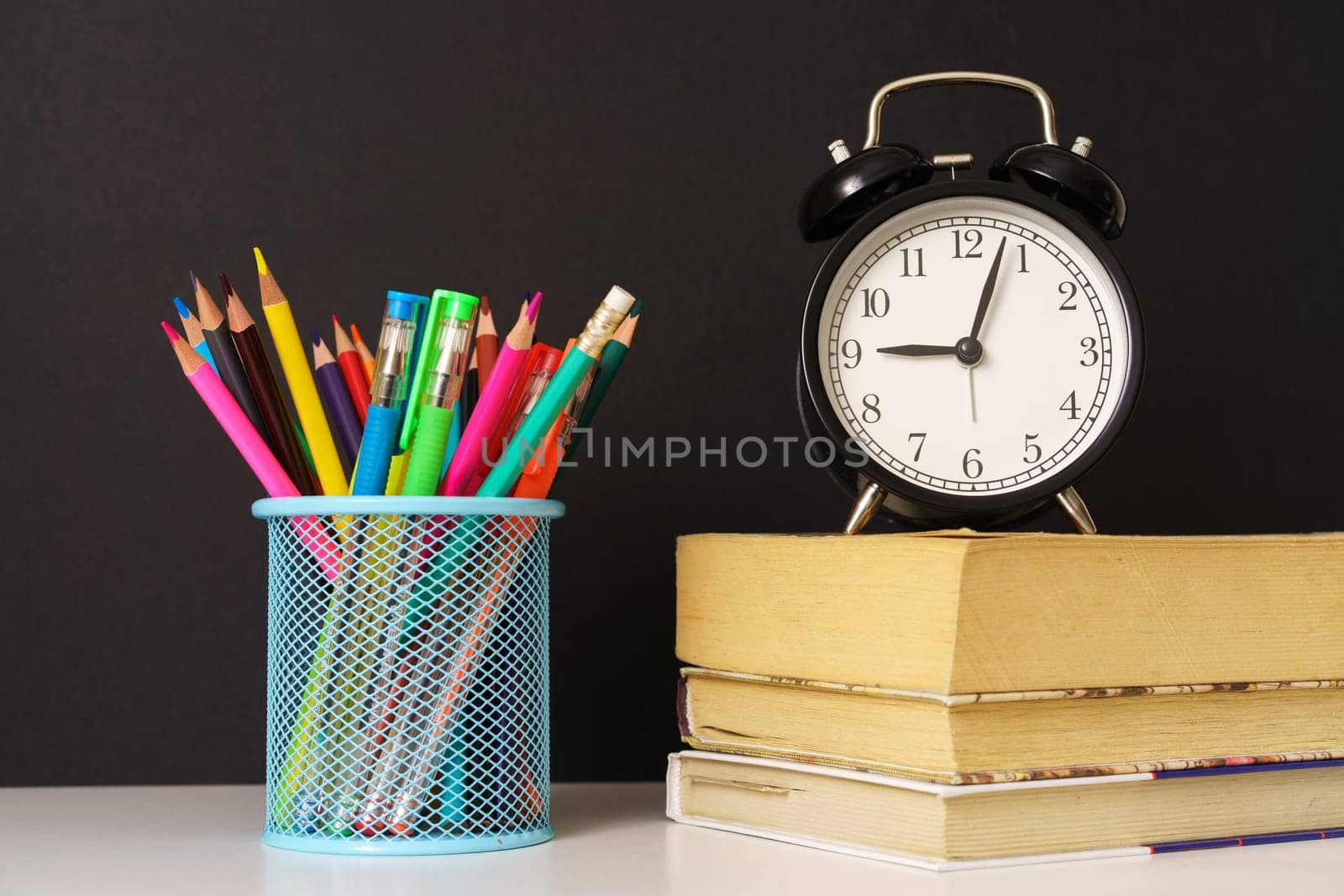 the alarm clock stands on the books and glass with pencils on black background, concept back to school