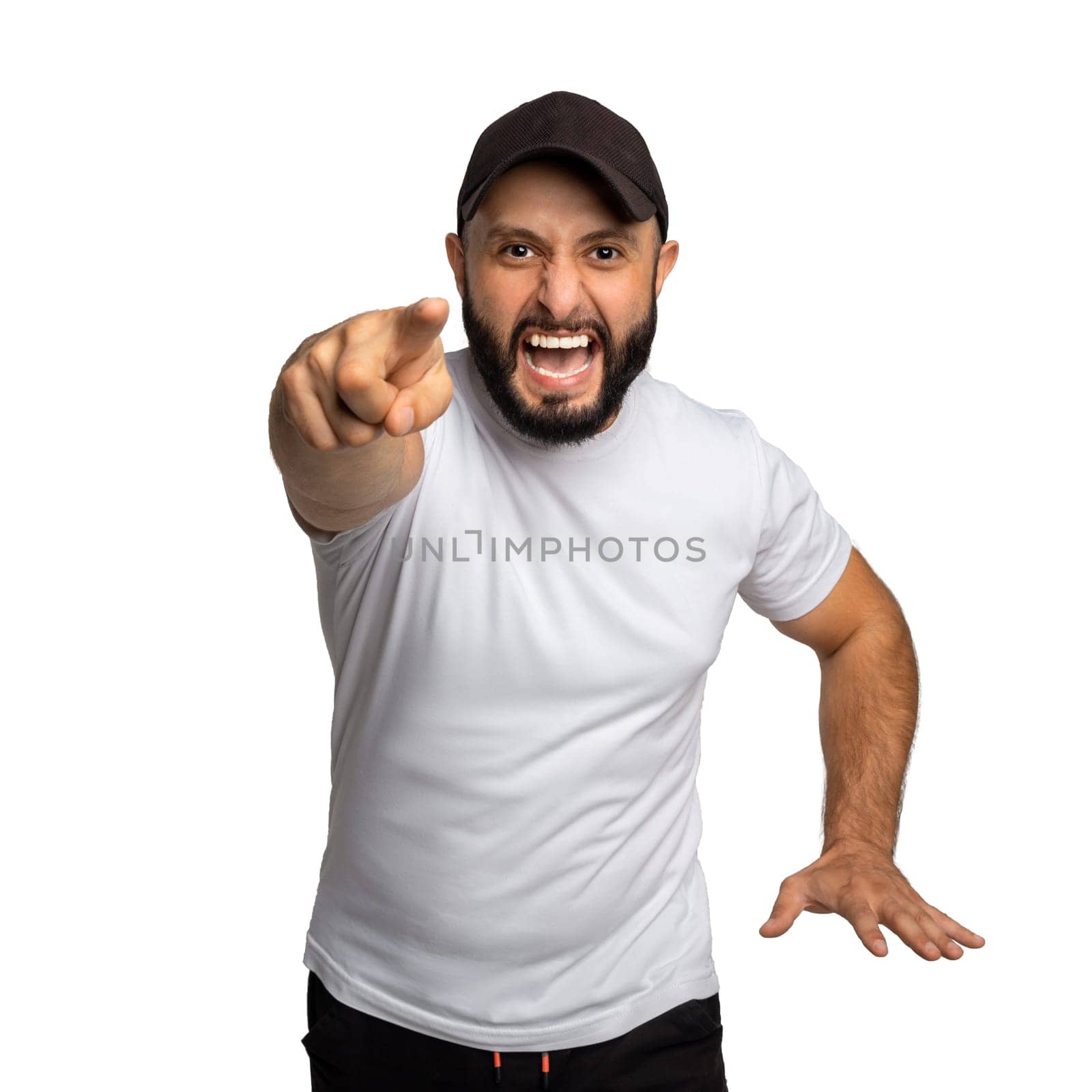 Portrait of peevish bearded guy with black hat, yells angrily at somebody, points with index finger directly at camera, dressed in white t shirt, blames you in doing wrong things. Indoor studio shot isolated on white background.
