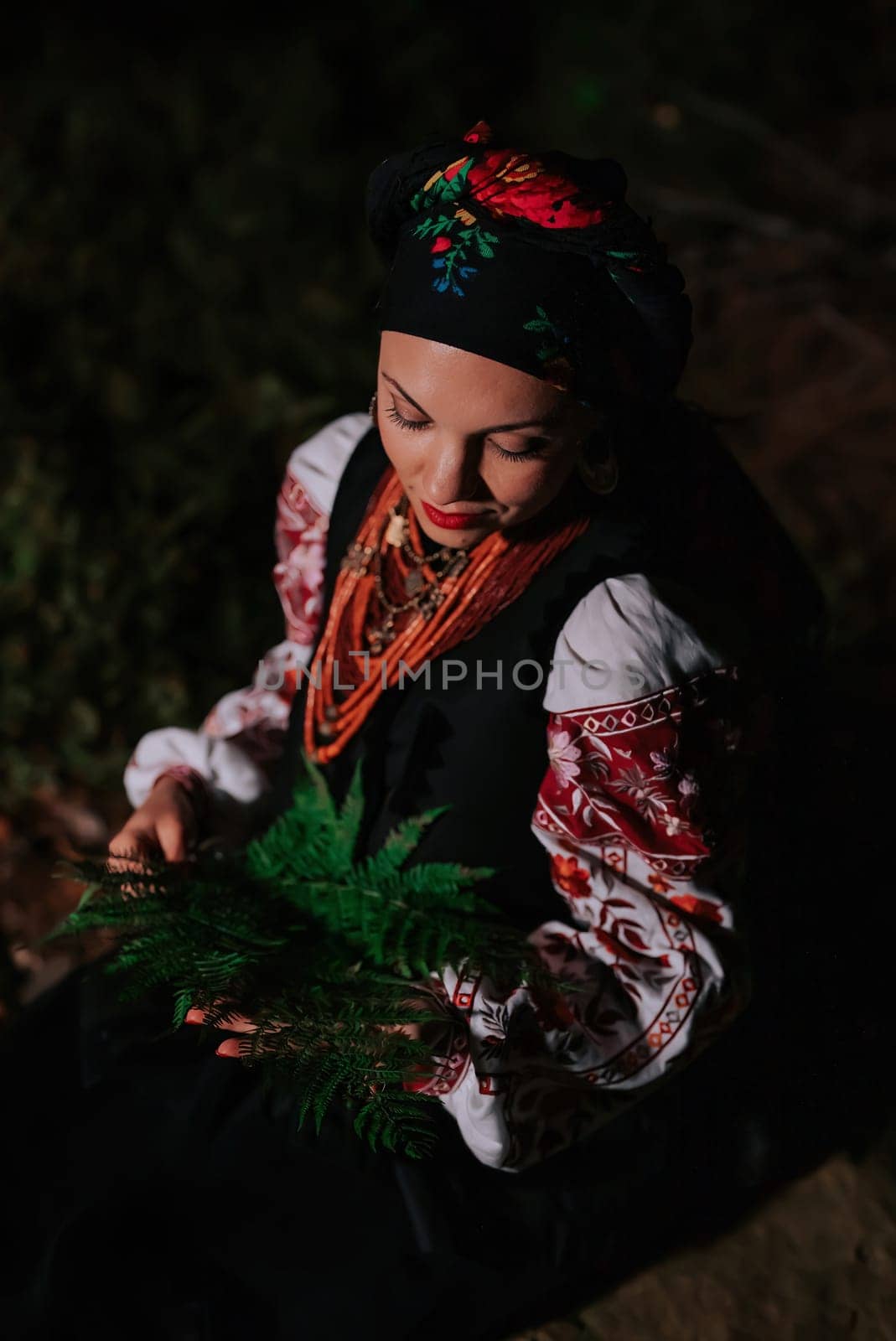 Witch woman collects herbs, ferns at night in Carpathian mountains forest. She in in traditional ukrainian handkerchief, national dress - vyshyvanka, ancient coral beads. Folk medicine concept.