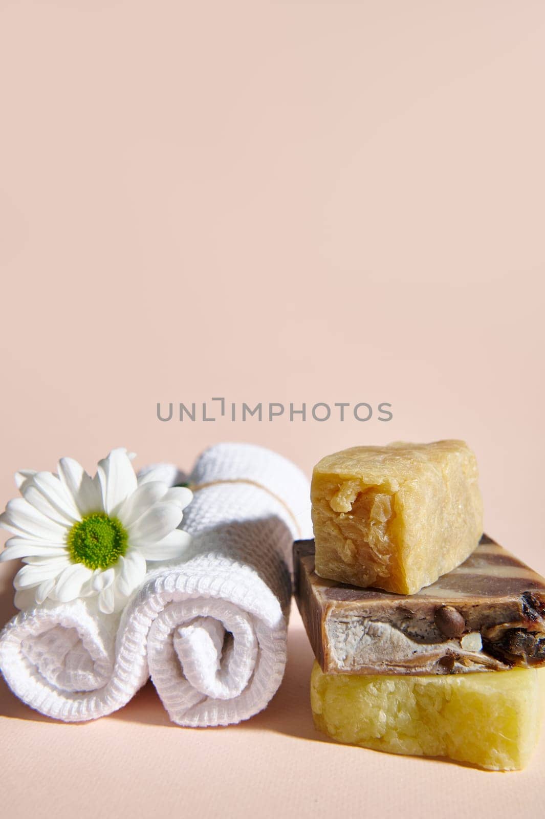 Spa set with two rolled white towels, chamomile flower and stack of organic natural soap bars with natural ingredients on isolated beige background. Vertical photo. Still life. Copy advertising space