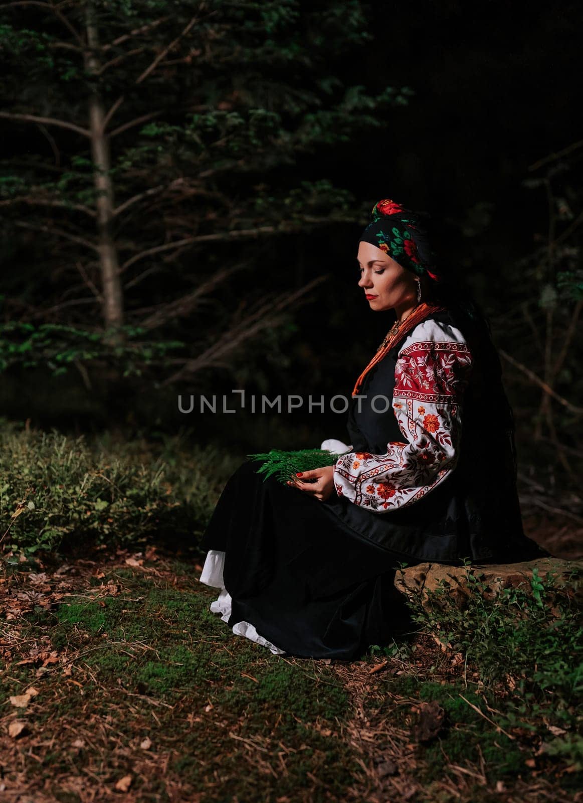 Witch woman collects herbs, ferns at night in Carpathian mountains forest. She in in traditional ukrainian handkerchief, national dress - vyshyvanka, ancient coral beads. Folk medicine concept.
