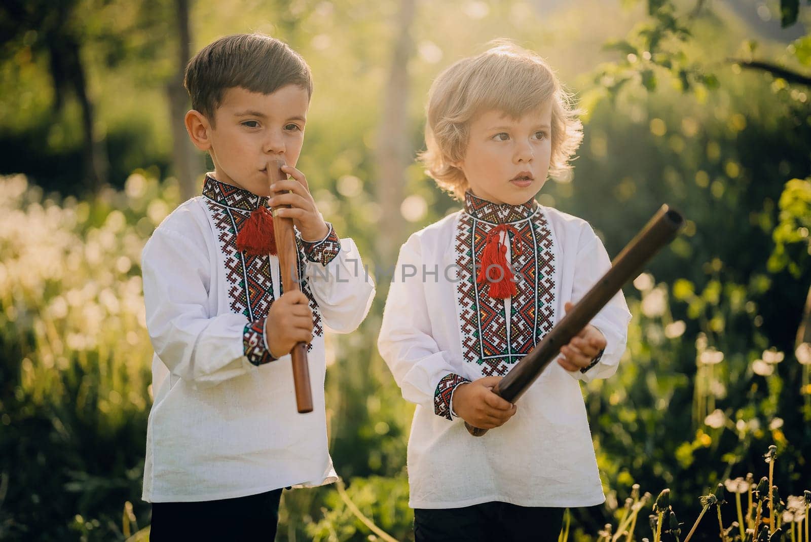 Little brothers boys playing on woodwind wooden flute - ukrainian sopilka outdoors. Folk music concept. Musical instrument. Children in traditional embroidered shirts - vyshyvanka.
