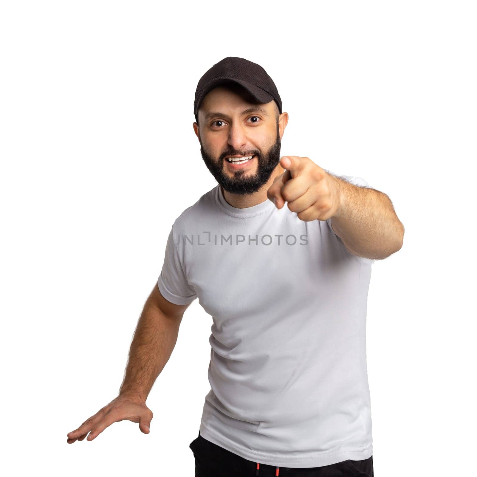 Portrait of peevish bearded guy with black hat, yells angrily at somebody, points with index finger directly at camera, dressed in white t shirt, blames you in doing wrong things. Indoor studio shot isolated on white background.