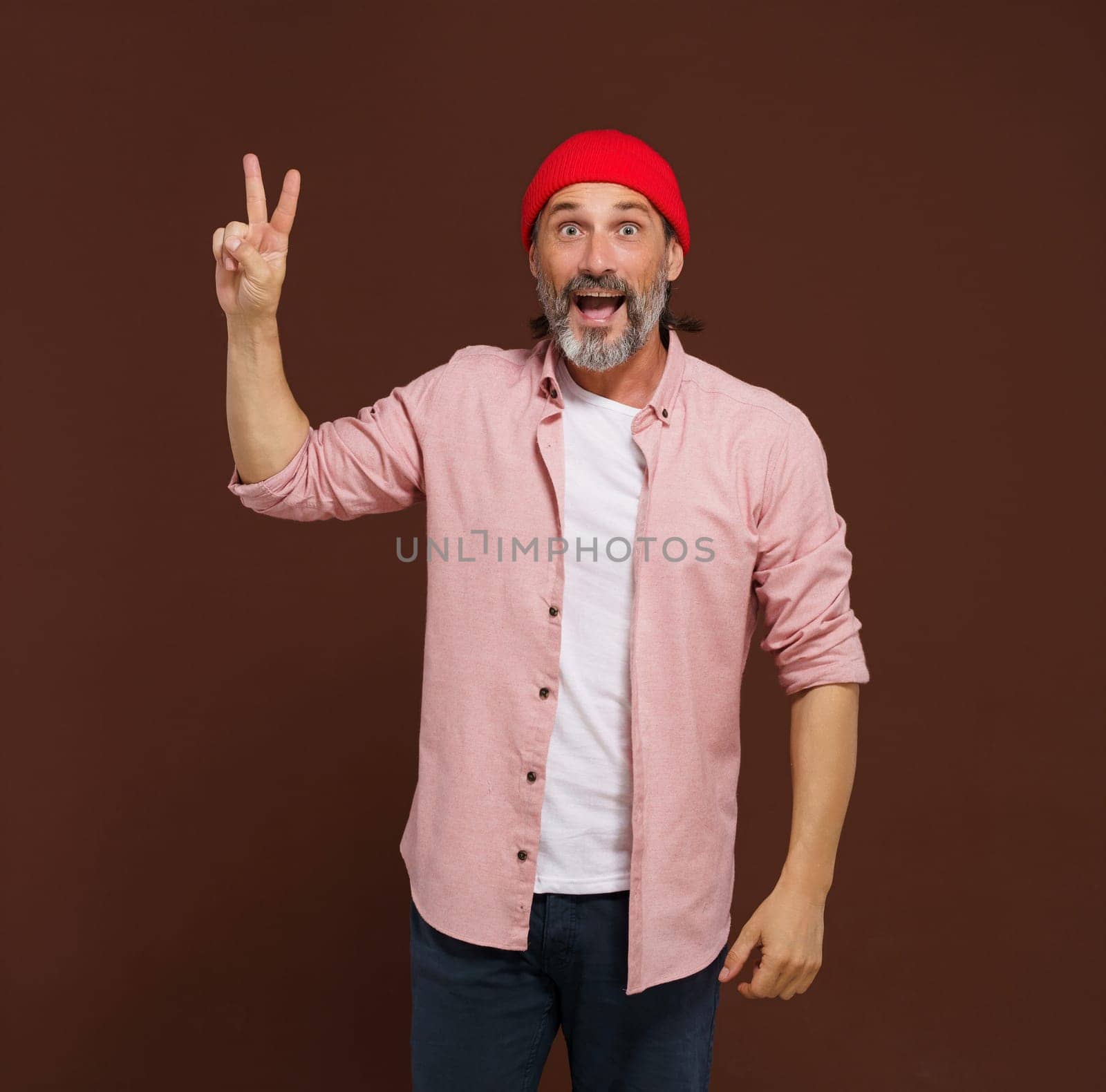 Positive European caucasian man handsome shows gesture of V victory. Cheerful man with silver beard raises his hand with two fingers V shape, indicating gesture of victory, with smile on face. by LipikStockMedia
