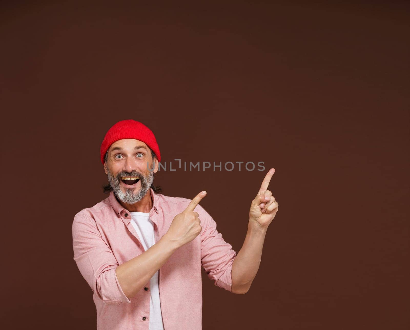 Amazing reaction. a mature European caucasian man points with both hands to the side with copy space isolated on brown. by LipikStockMedia