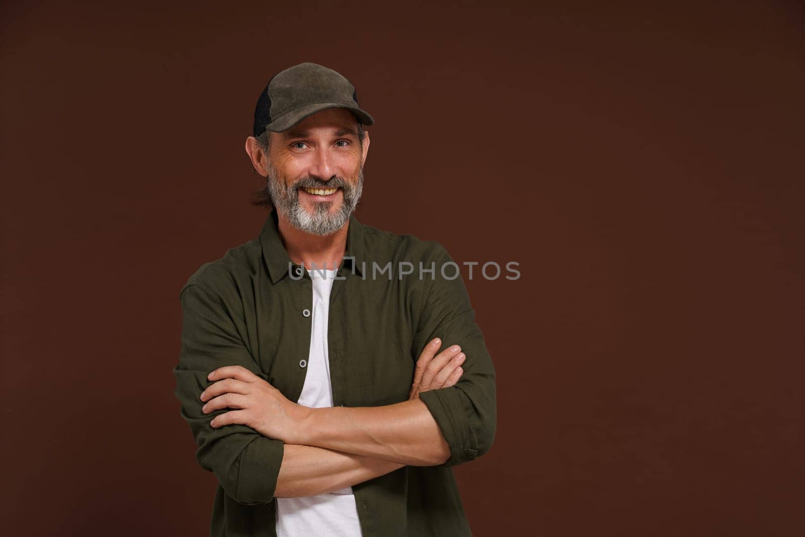 Happy Smiling man. A fisherman in a baseball cap and green shirt stands cross-legged against a brown background with copy space. by LipikStockMedia