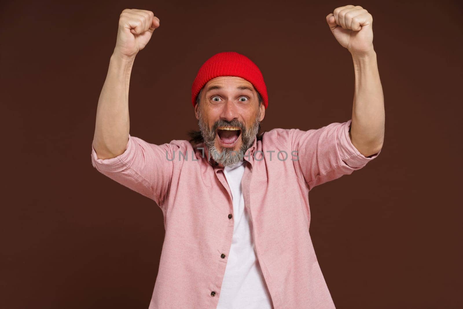Sports fan victory concept. Happy mature handsome European caucasian man holds clenched fists above his head celebrating win of his team command. by LipikStockMedia