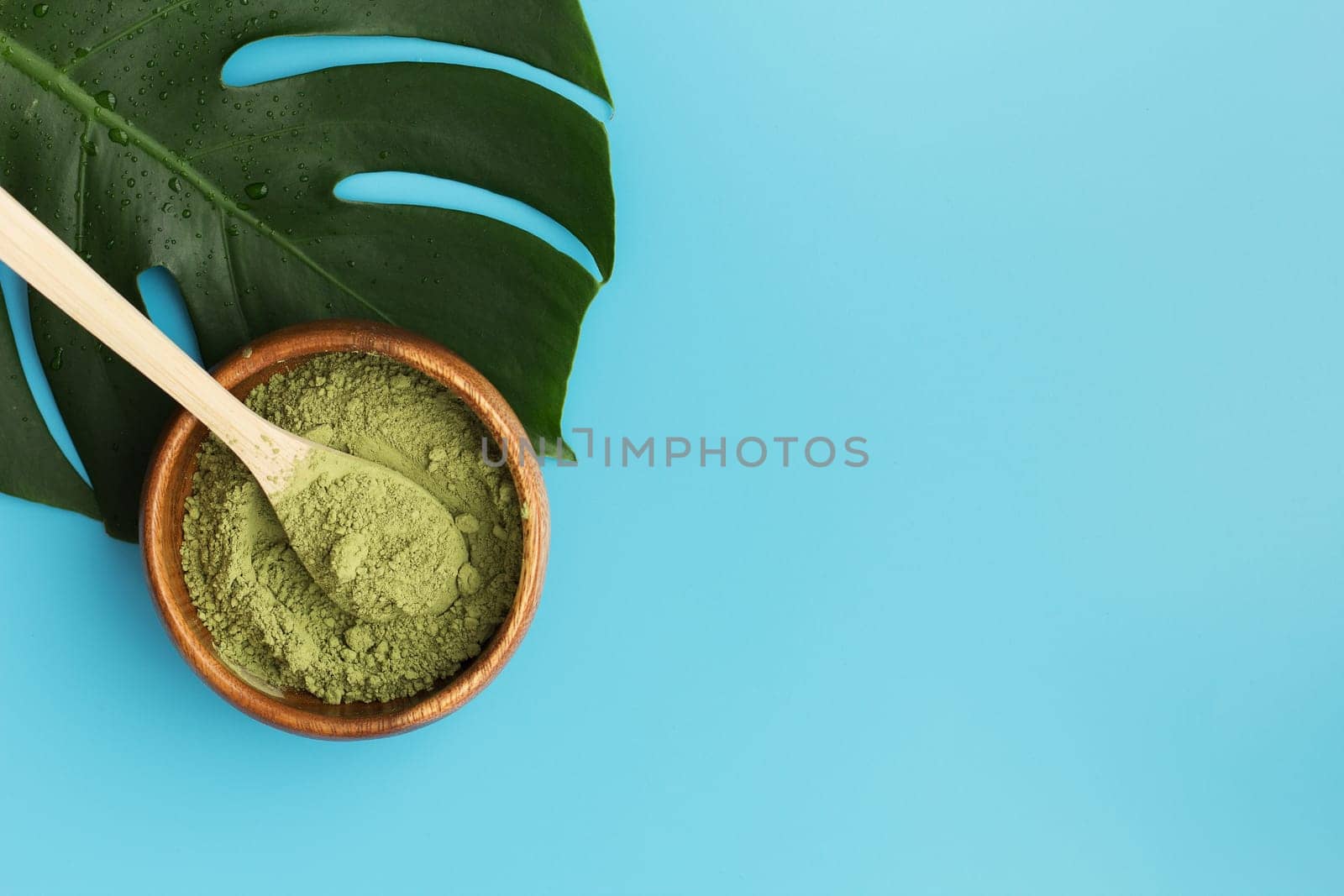 Wooden spoon with matcha tea in a wooden plate and monstera leaf on a blue background. Matcha green tea powder. copy space