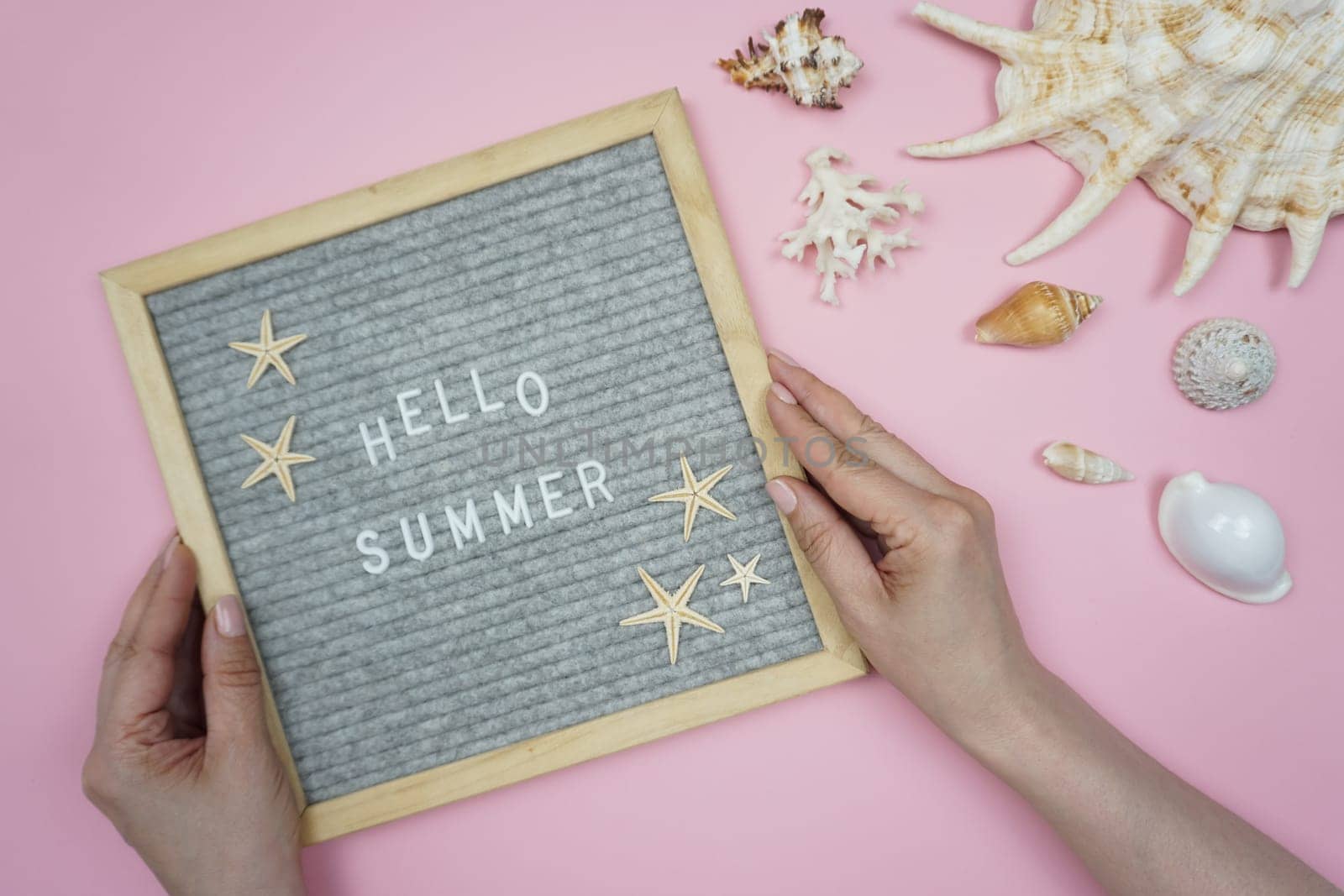 A woman holds a board with the inscription Hello, summer in her hands. On a pink background are a variety of shells.