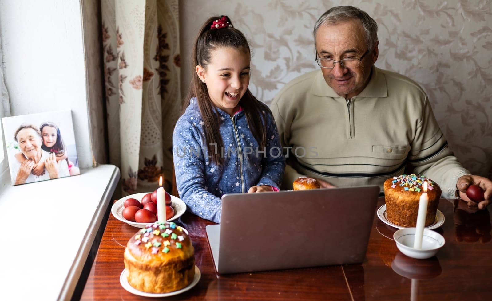 grandfather and granddaughter are talking via video link to their friends. Decorated table with colorful eggs and cake. Chatting during the COVID pandemic and the Easter holidays by Andelov13