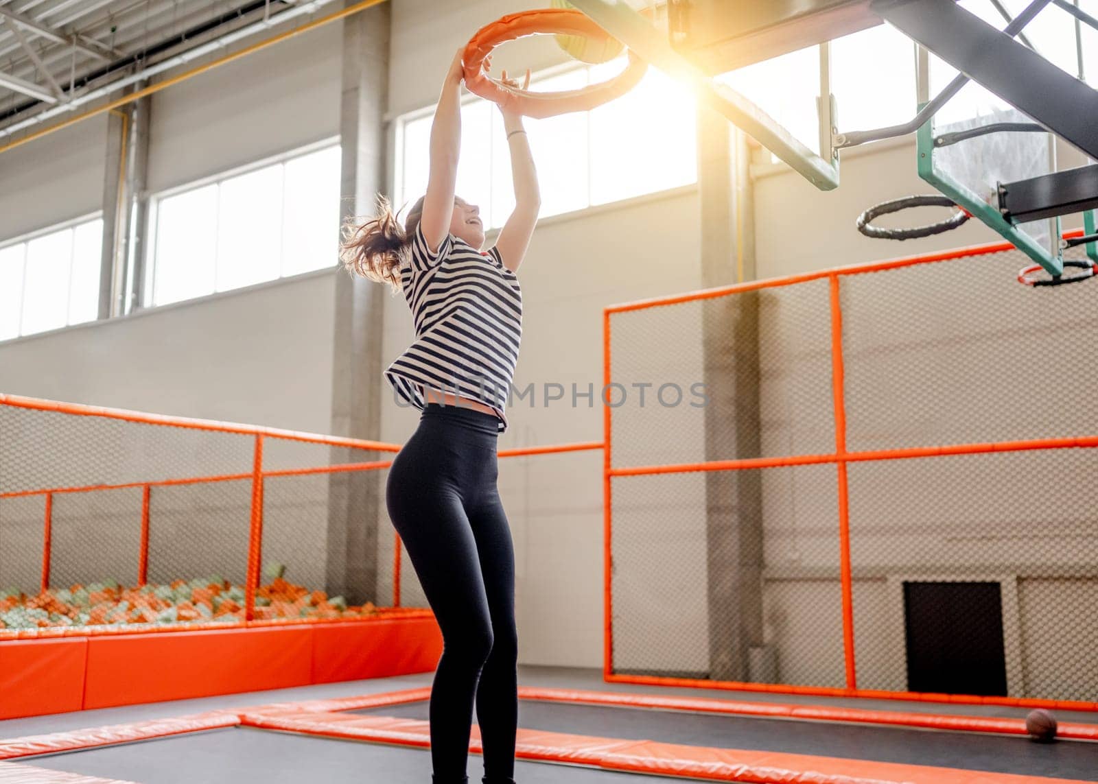 Pretty girl in trampoline park jumping and holding basketball basket. Happy teenager enjoying amusement activities