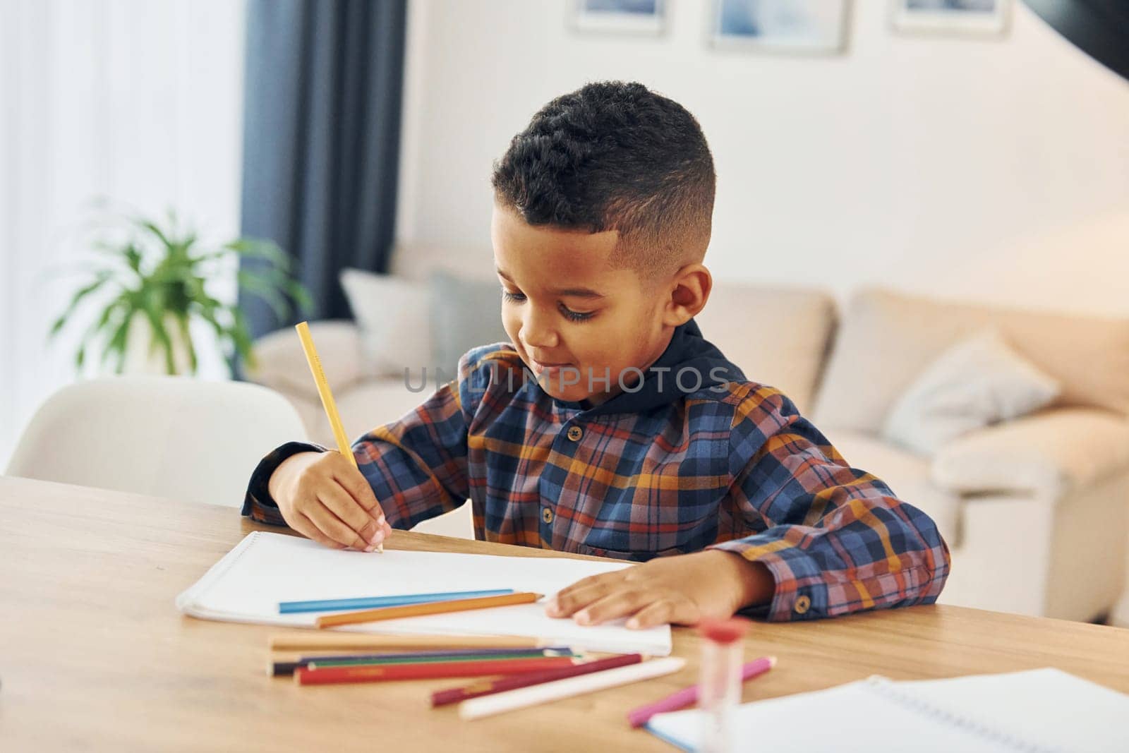 Sitting by the table. Cheerful young boy is at home alone.
