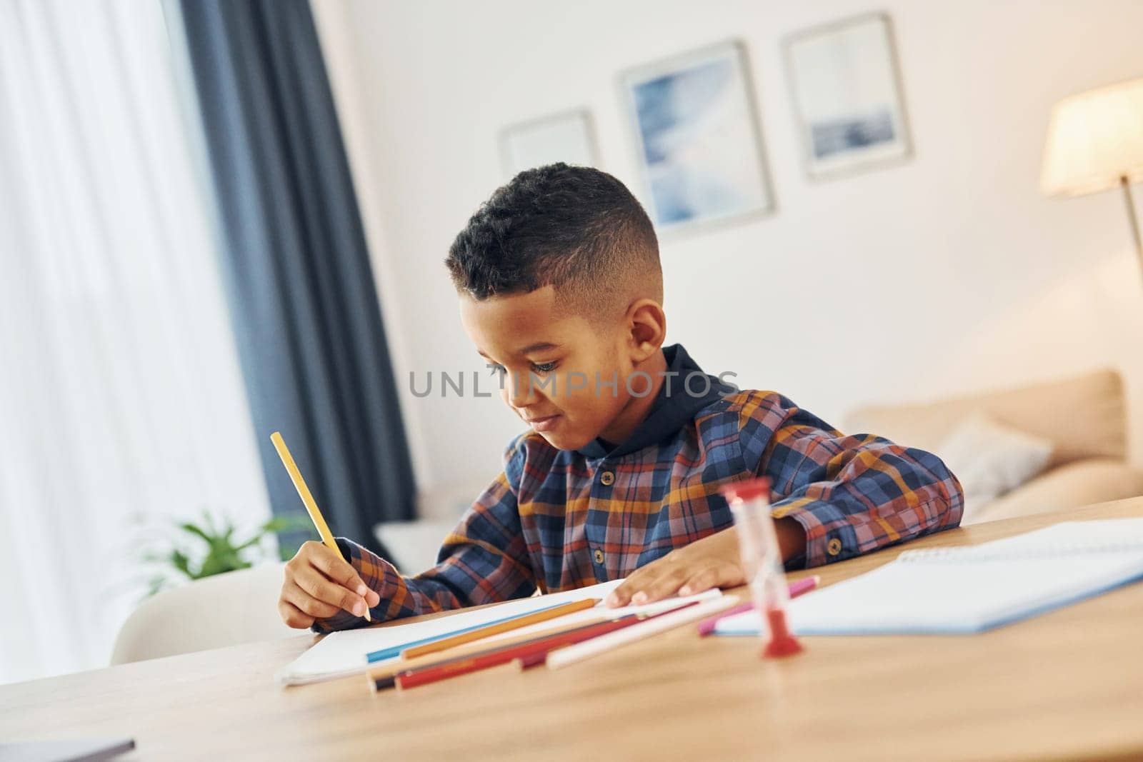Sitting by the table. Cheerful young boy is at home alone.