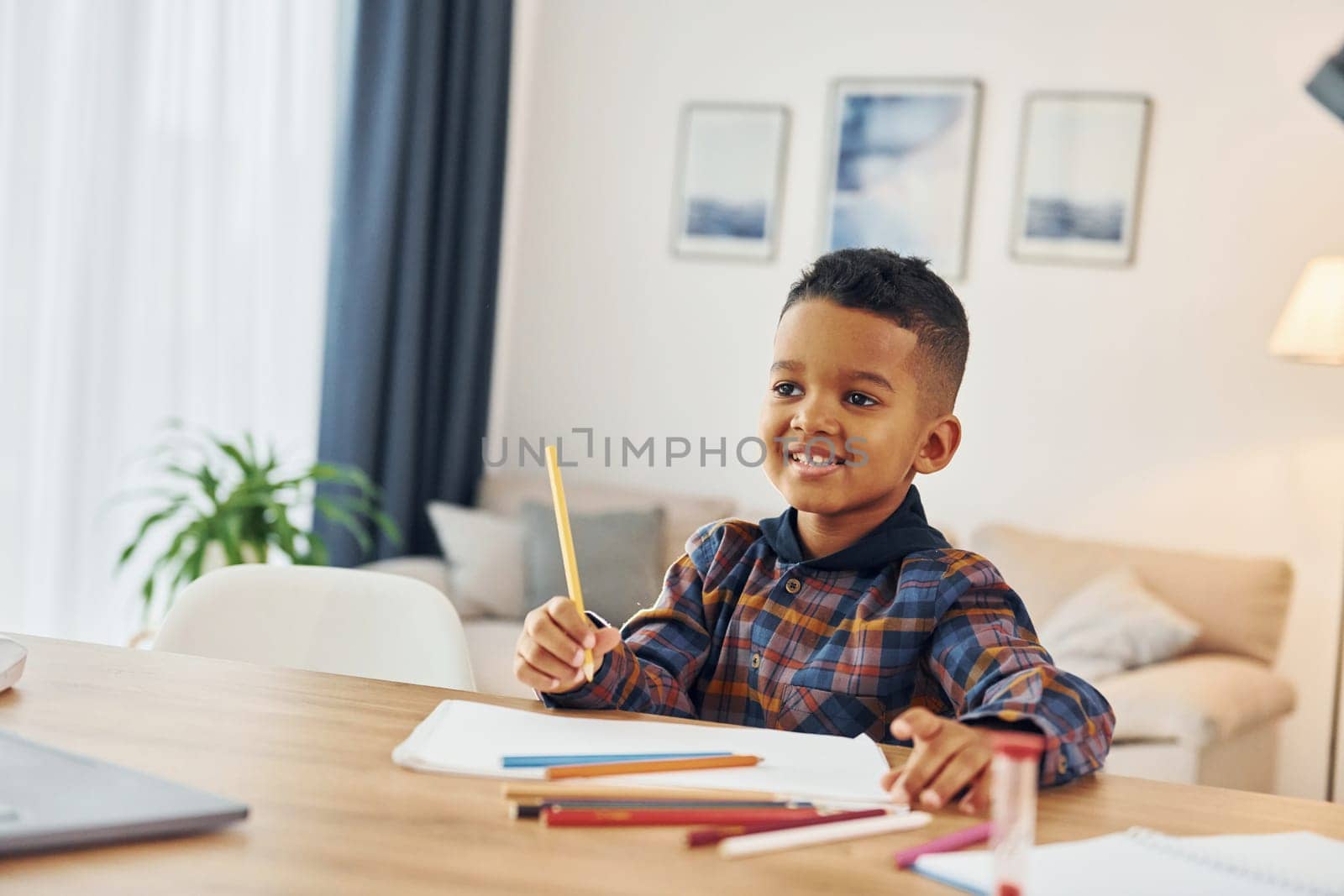 Sitting by the table. Cheerful young boy is at home alone.