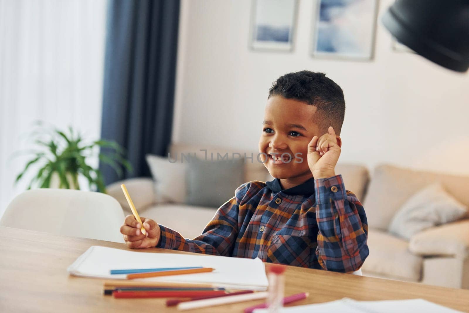 Sitting by the table. Cheerful young boy is at home alone.