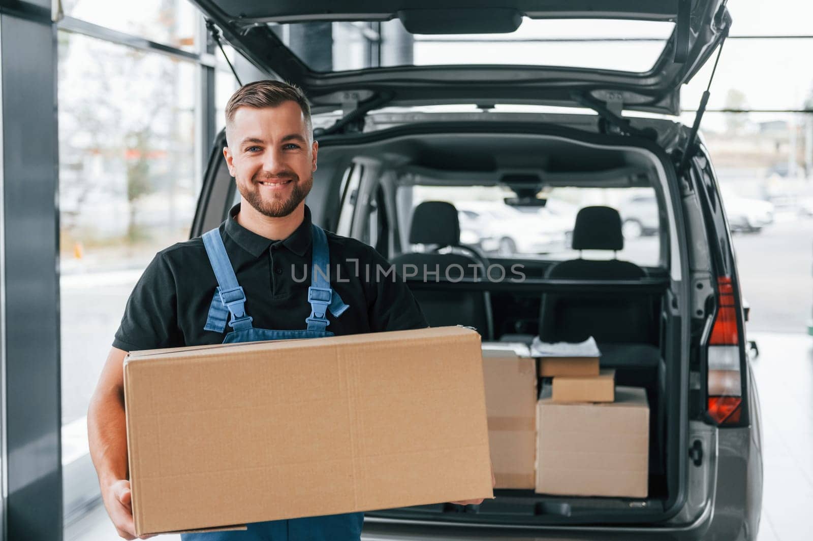 Holding box. Delivery man in uniform is indoors with car and with order.