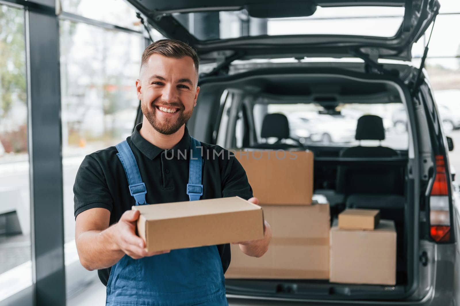 Holding box. Delivery man in uniform is indoors with car and with order.