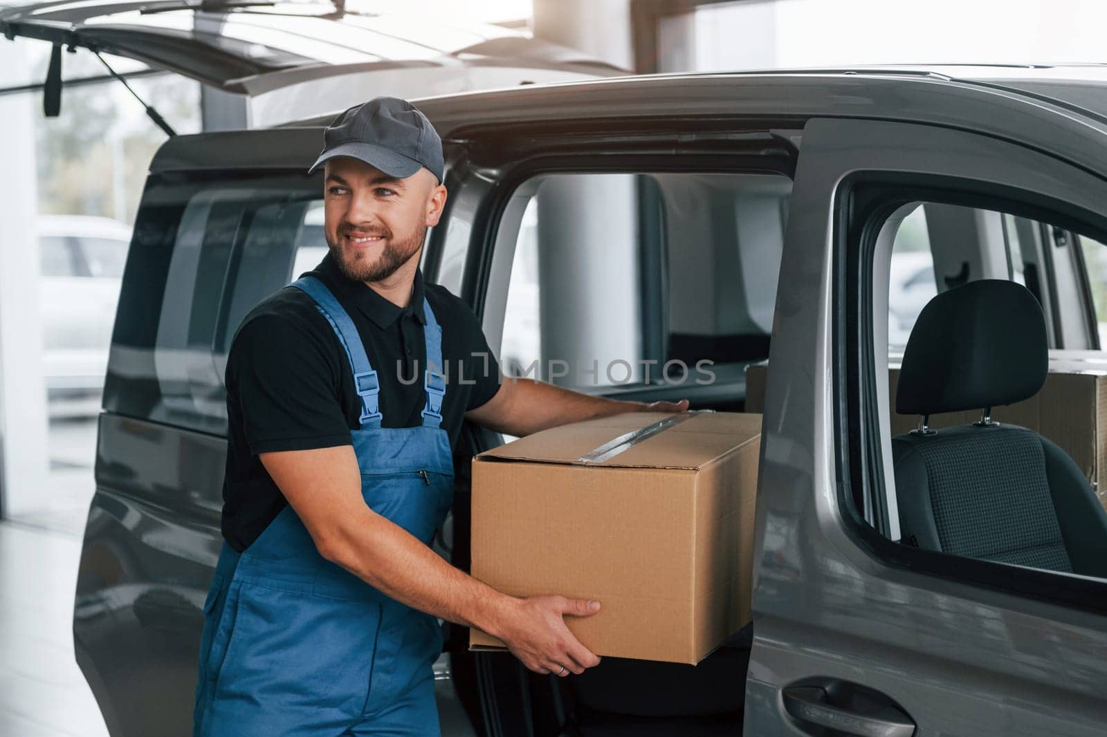 With box in hands. Delivery man in uniform is indoors with car and with order.