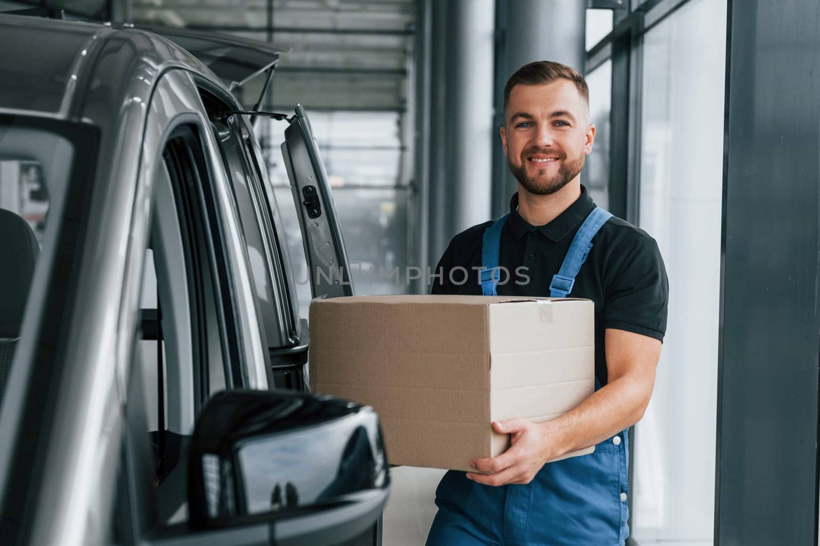 With box in hands. Delivery man in uniform is indoors with car and with order.