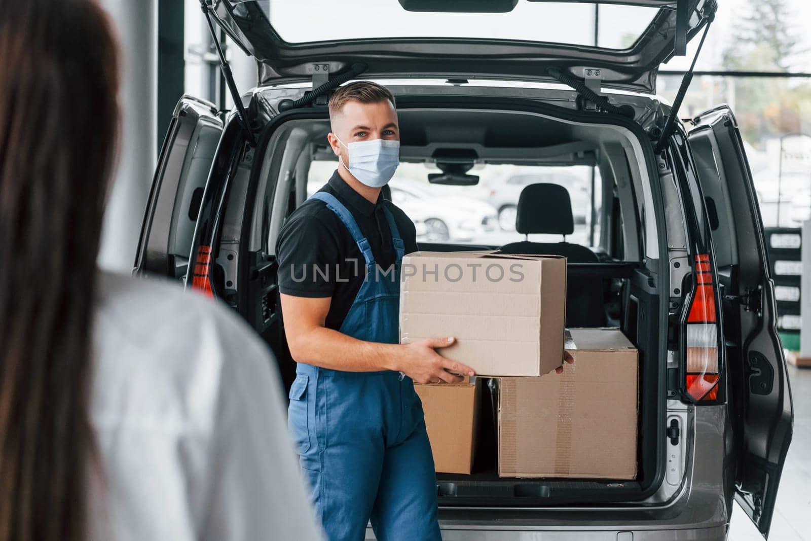 Woman waits for her order. Delivery man in uniform is indoors with car.