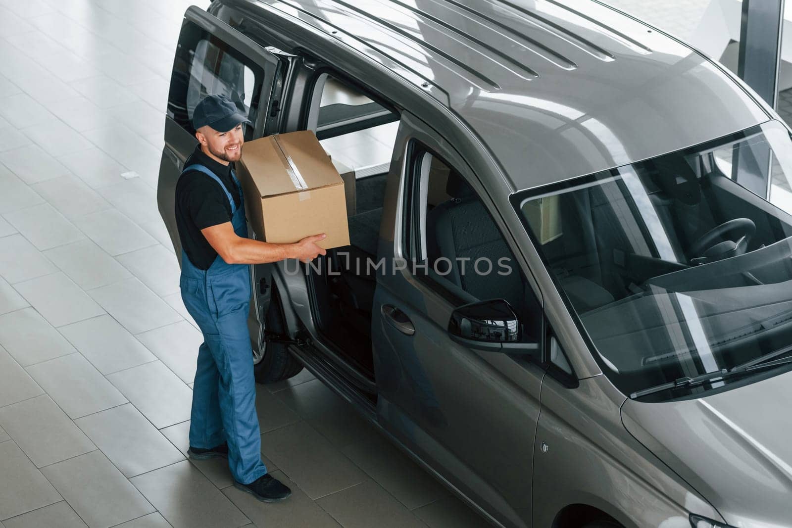 In blue colored clothes. Delivery man in uniform is indoors with car and with order.